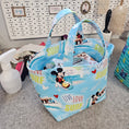 Load image into Gallery viewer, A light blue fabric tote bag with a Mickey Mouse and Minnie Mouse surf-themed print, featuring the words 'SURF', 'LOVE', and 'Mickey', shown on a gray carpeted floor next to a white pegboard.
