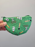 Load image into Gallery viewer, A hand holding a green zippered pouch with a printed pattern of scissors, paintbrushes, and rockets on a plain gray background.