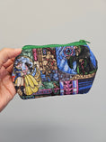 Load image into Gallery viewer, A hand holding a small zippered pouch with a colorful stained-glass-style print featuring Belle, a wizard, and other fantasy characters, against a plain light gray background.