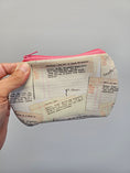 Load image into Gallery viewer, A hand holding a small zippered pouch with a pink zipper and a cream-colored fabric printed with vintage fashion magazine page designs featuring text and grid lines.