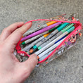 Load image into Gallery viewer, Hand holding a pencil case with pens and markers on a concrete surface