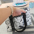 Load image into Gallery viewer, Hand holding a patterned bag with black straps on a tiled floor background