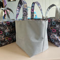 Load image into Gallery viewer, A light gray fabric tote bag with dark patterned handles and a floral print body, displayed on a table with two matching bags partially visible in the background.