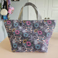Load image into Gallery viewer, A tote bag with a dark gray and black galaxy print, decorated with colorful daisy-like flowers and banners reading 'I CAN BUY MYSELF FLOWERS', sitting on a table.