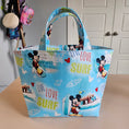 Load image into Gallery viewer, A light blue fabric tote bag with Mickey Mouse and Minnie Mouse prints, featuring 'LIFE LOVE SURF' text and surfboard motifs, shown hanging against a beige wall.