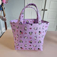 Load image into Gallery viewer, A light purple fabric tote bag with a repeating cartoon Minnie Mouse and Mickey Mouse pattern, hanging against a beige wall next to white paneled cabinets.