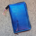 Load image into Gallery viewer, A metallic blue zip-around pouch with a subtle iridescent sheen, resting on a gray textured carpet. The pouch has black trim and a black zipper pull, with a small retail tag attached near the bottom left.