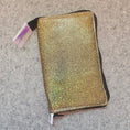 Load image into Gallery viewer, A rectangular metallic gold glitter pouch with black trim and a white zipper pull, lying on a gray felt surface.