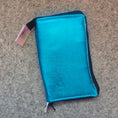 Load image into Gallery viewer, A metallic turquoise blue zippered pouch with black trim, photographed on a gray felt surface.