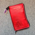 Load image into Gallery viewer, A metallic red zippered pouch or wallet with black trim, shown on a gray textured carpet with a small retail tag attached.