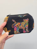 Load image into Gallery viewer, A hand holding a small zippered pouch with a black background and a colorful, patterned jaguar design. The pouch features a black base with a vibrant, multicolored jaguar motif outlined in gold, set against a plain light gray background.