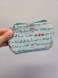 Load image into Gallery viewer, A hand holding a small zippered pouch with a light blue background printed with black text, red hearts, and small cute illustrations like a panda face, flowers, cupcakes, and a coffee cup, against a plain light gray wall.