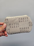 Load image into Gallery viewer, A hand holding a small zippered pouch made of light beige fabric printed with the word 'MAKER maker' in black text, against a plain gray background.