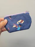 Load image into Gallery viewer, A hand holding a small zippered pouch with a blue tropical leaf pattern and a cartoon character on the front, set against a plain light gray background.