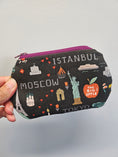 Load image into Gallery viewer, A hand holding a small zippered pouch with a black background printed with travel-themed illustrations and text including Istanbul, Moscow, Tokyo, The Big Apple, and The Statue of Liberty. The pouch has a purple zipper and is photographed against a plain light gray background.