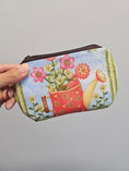 Load image into Gallery viewer, A hand holding a small zippered pouch with a colorful gardening print featuring a red watering can, pink flowers, green leaves, and a light blue background.