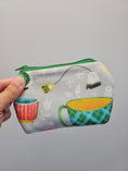 Load image into Gallery viewer, A hand holding a small zippered pouch with a colorful tea-themed print, featuring illustrated teacups and hanging tea bags on a light gray background.