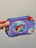 Load image into Gallery viewer, A hand holding a small zippered pouch with a purple background and Disney's The Little Mermaid character print, shown against a plain light gray background.
