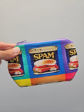 Load image into Gallery viewer, A hand holding a small zippered pouch with a colorful Spam tin print pattern against a plain gray background.