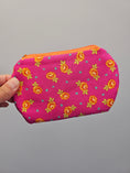 Load image into Gallery viewer, A hand holding a small zippered pouch with a bright pink background and an orange floral print, set against a plain gray background.