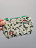Load image into Gallery viewer, A hand holding a small zippered pouch with a white background and a green holly and ornament print, featuring a teal zipper.