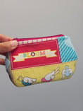 Load image into Gallery viewer, A hand holding a small zippered pouch with a colorful patchwork fabric design featuring the word 'BLOOM' on a red panel and cartoon birds on a yellow panel, against a plain gray background.
