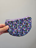 Load image into Gallery viewer, A hand holding a small zippered pouch with a dense floral print of purple, blue, and white flowers with green leaves on a black background, set against a plain light gray wall.