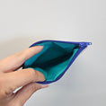 Load image into Gallery viewer, Hand holding a teal pouch with a purple zipper against a light gray background
