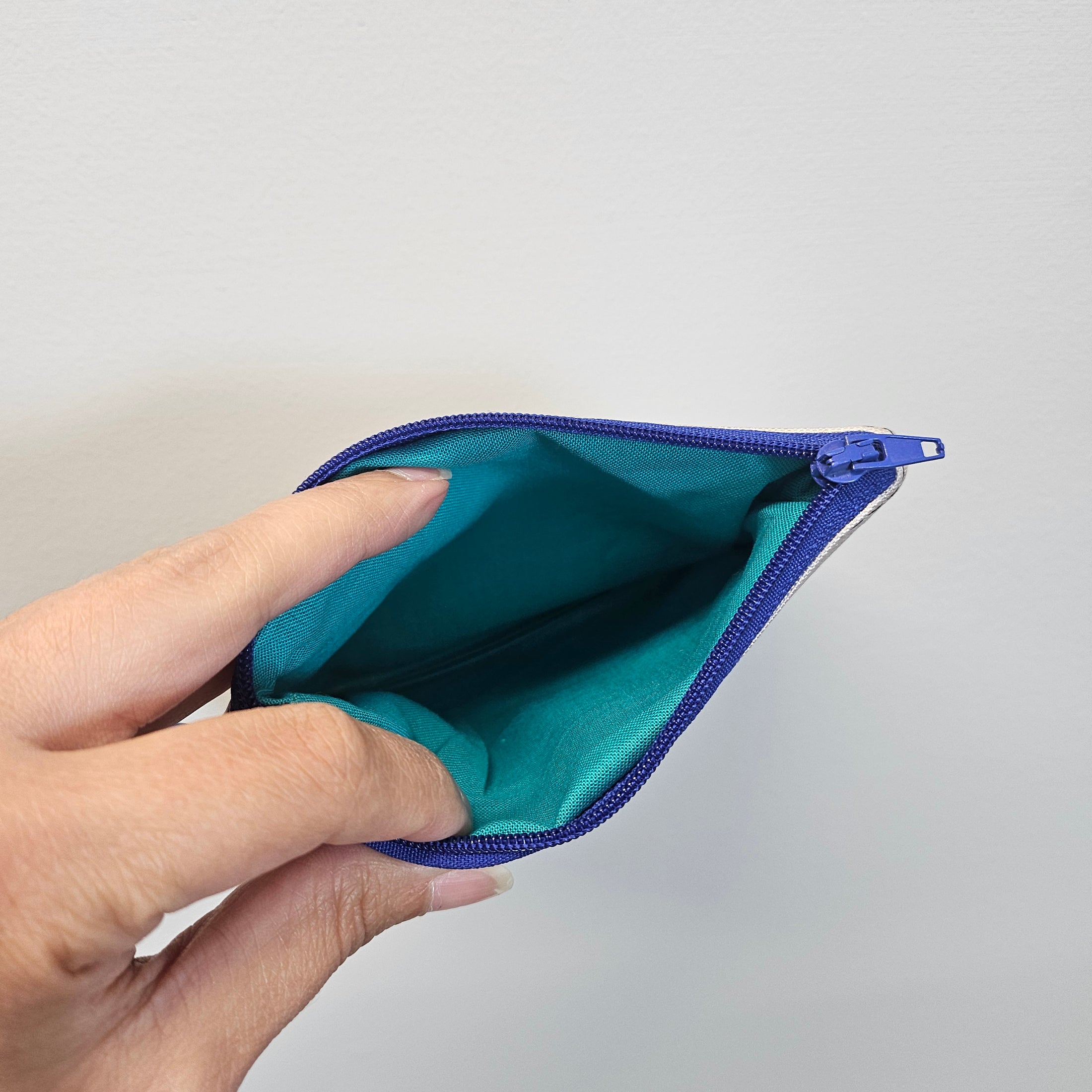 Hand holding a teal pouch with a purple zipper against a light gray background