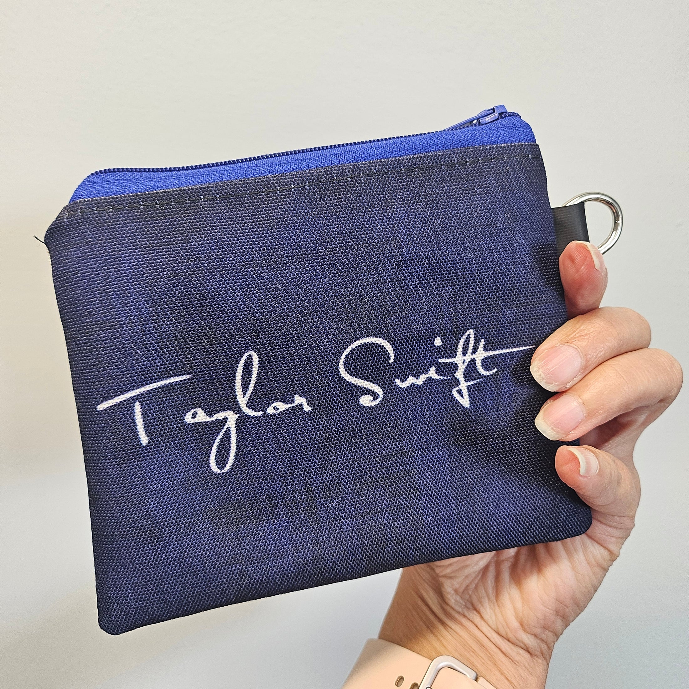 Hand holding a navy blue pouch with 'Taylor Swift' branding on a light gray background