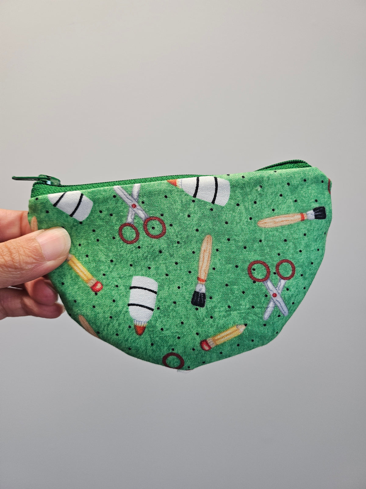 A hand holding a green zippered pouch with a printed pattern of scissors, paintbrushes, and rockets on a plain gray background.