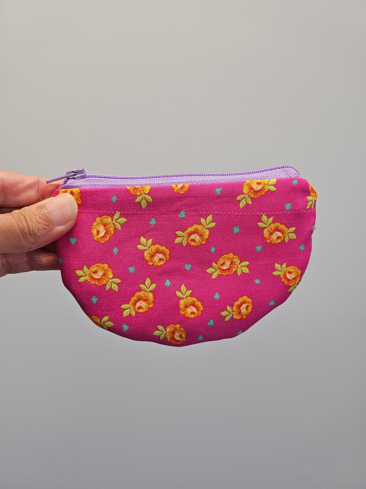 A hand holding a small fabric zip pouch with a bright pink background and an orange floral pattern, set against a plain gray background.