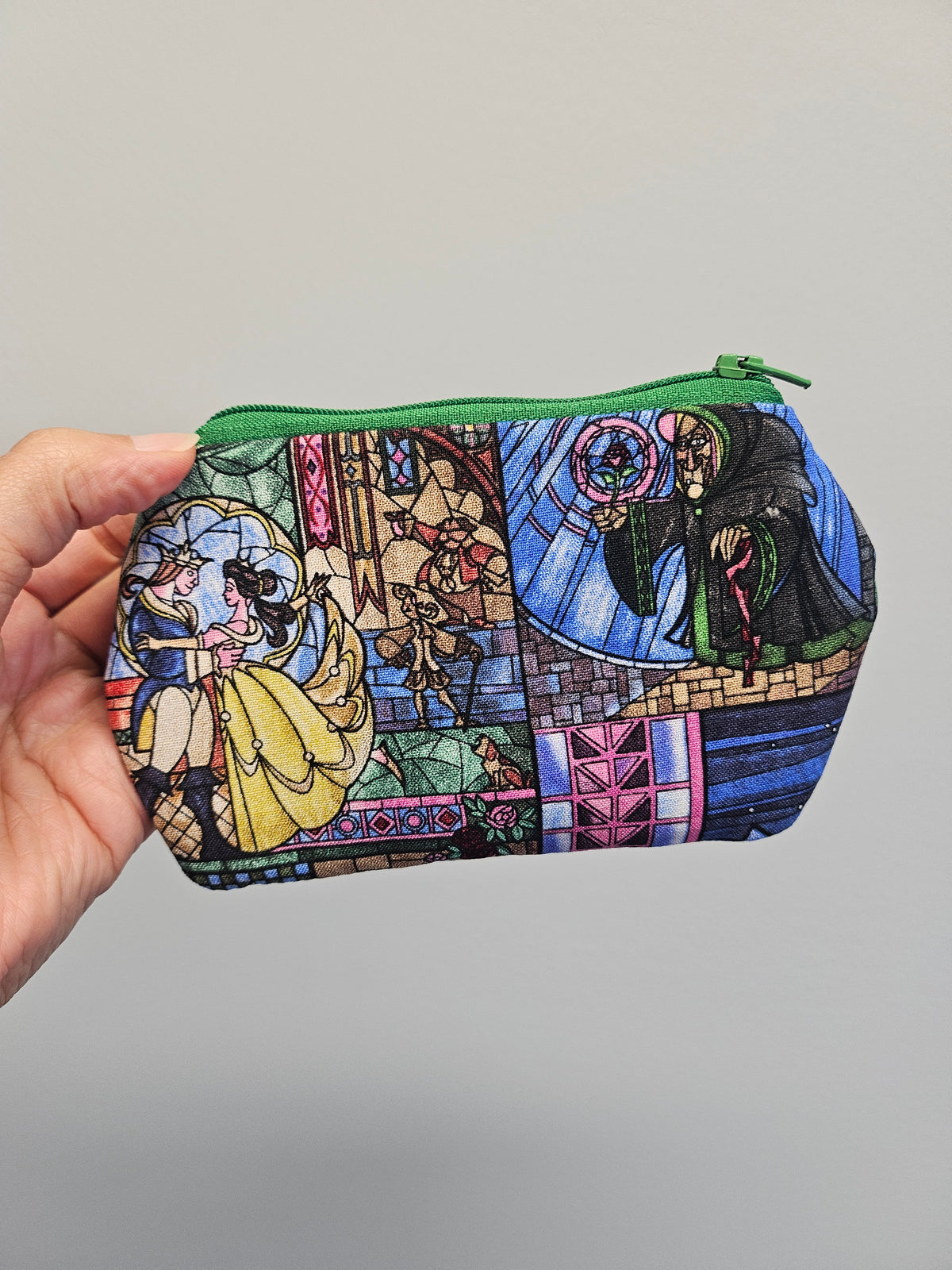 A hand holding a small zippered pouch with a colorful stained-glass-style print featuring Belle, a wizard, and other fantasy characters, against a plain light gray background.