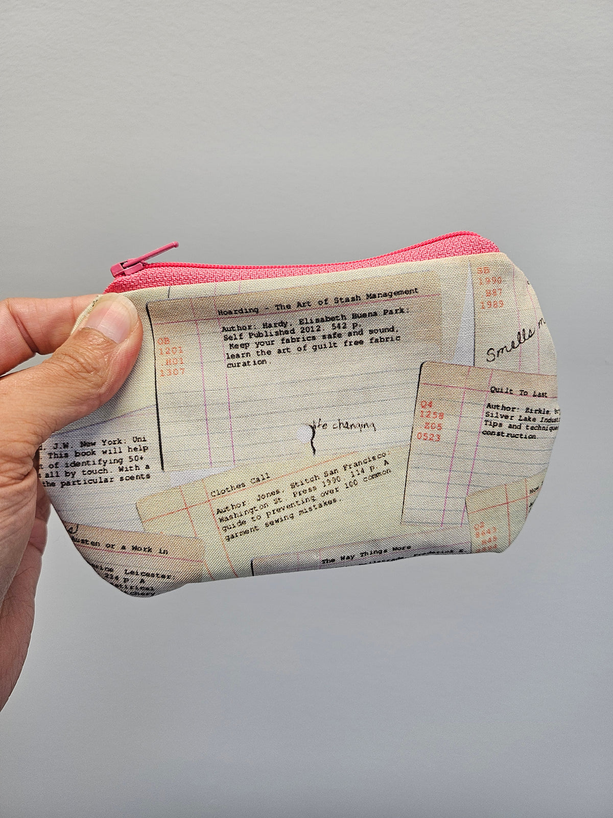 A hand holding a small zippered pouch with a pink zipper and a cream-colored fabric printed with vintage fashion magazine page designs featuring text and grid lines.