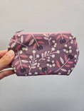 Load image into Gallery viewer, A hand holding a small zippered pouch with a floral pattern in dusty purple, pink, and white against a plain light gray background.
