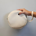 Load image into Gallery viewer, Hand holding a round beige clutch bag against a plain background

