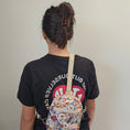 Load image into Gallery viewer, Person wearing a black t-shirt with text and a floral-patterned bag on a plain background
