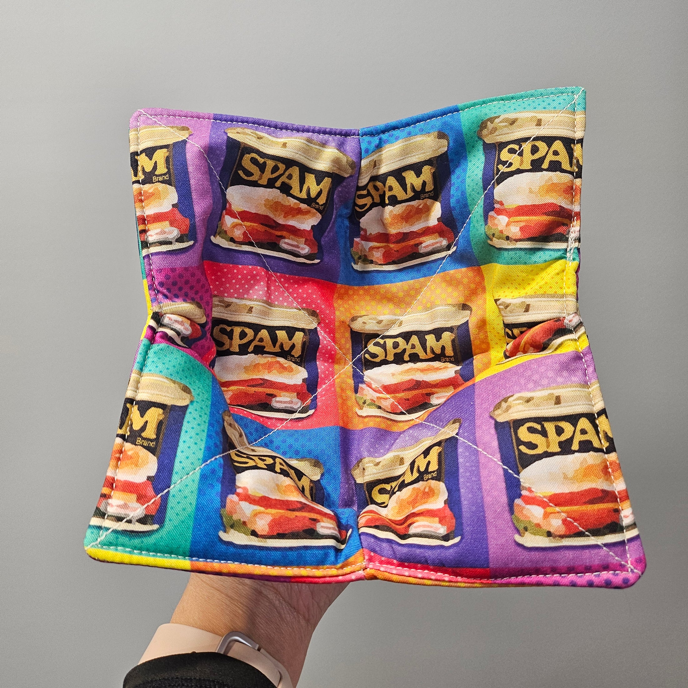 Colorful pot holder with Spam can pattern held by a hand against a gray background