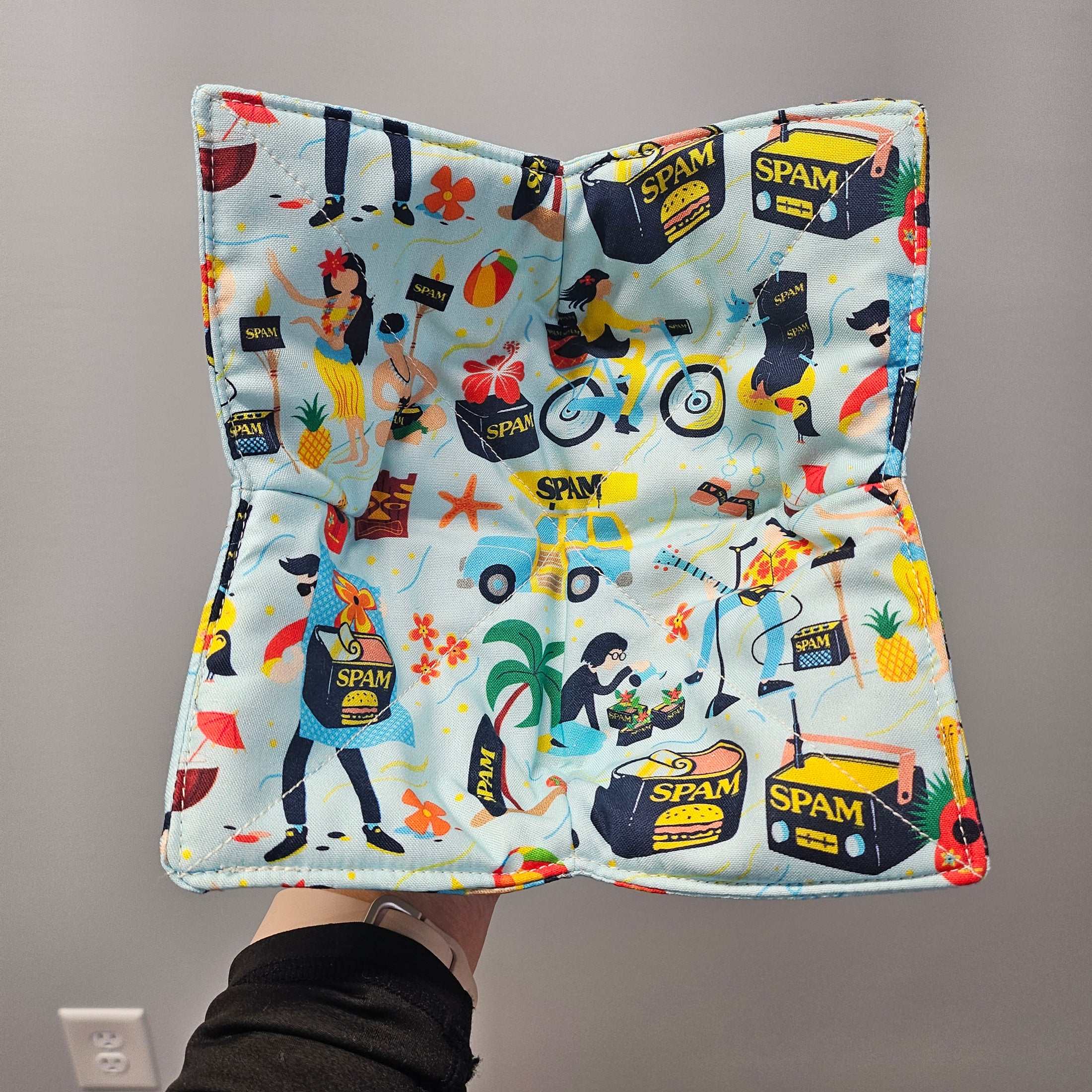 Colorful fabric with 'SPAM' logo and tropical design held by a person against a gray background