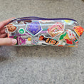 Load image into Gallery viewer, Clear plastic case with colorful stickers held by a hand on a gray background
