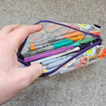 Load image into Gallery viewer, Hand holding a colorful pencil case with pens and markers on a gray surface
