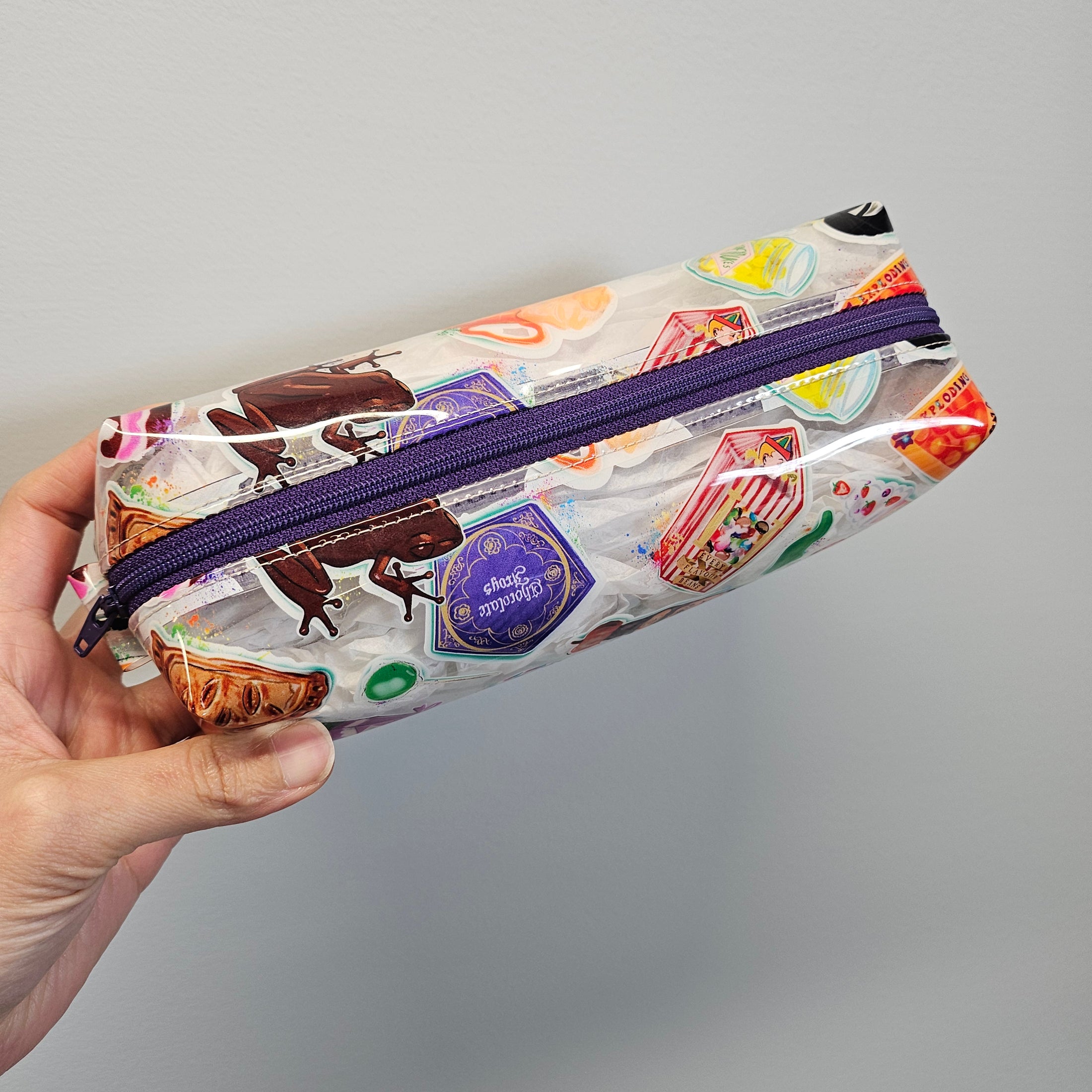 Clear pouch with colorful designs and a purple zipper held by a hand against a gray background