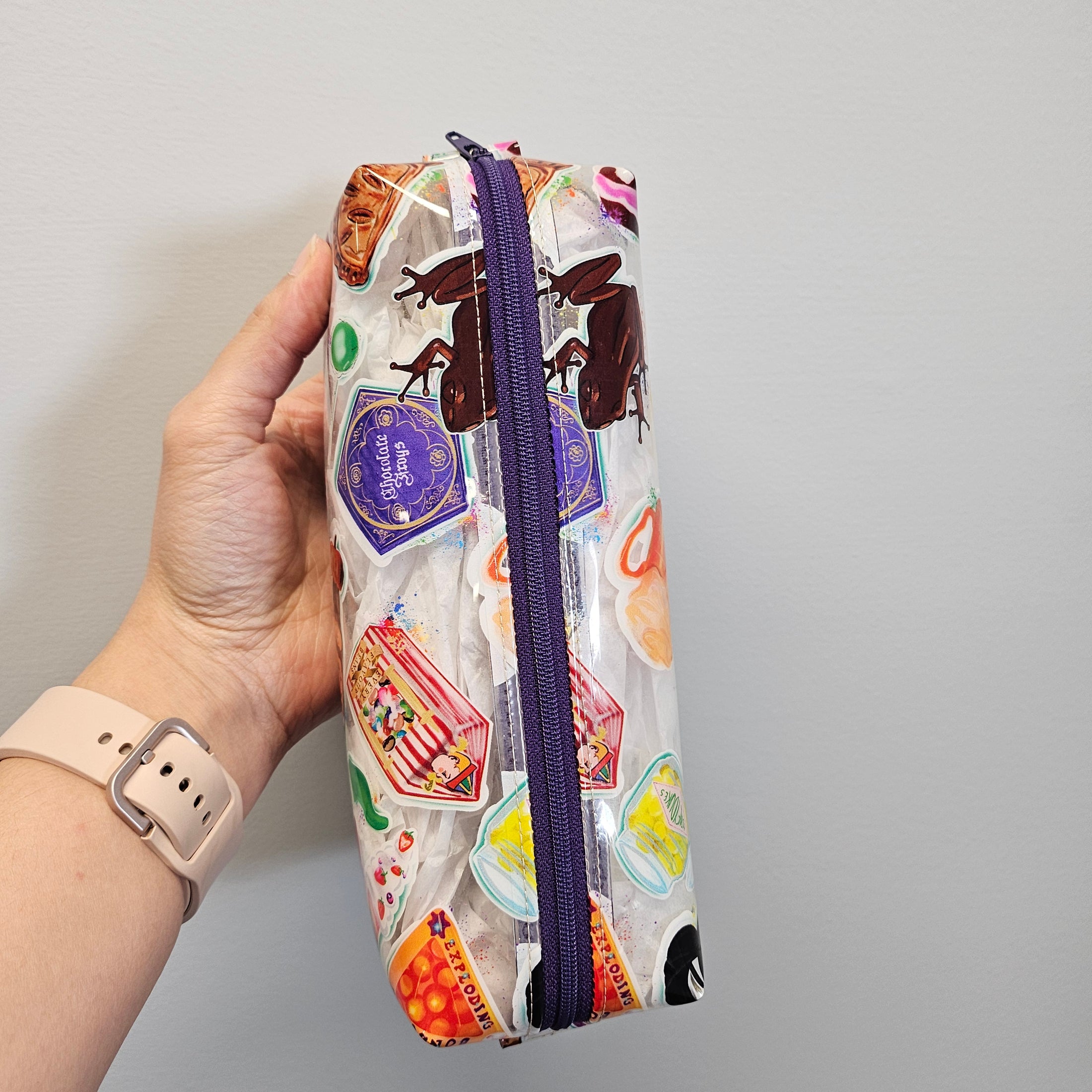 Hand holding a colorful pencil case with a purple zipper against a gray background