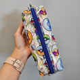 Load image into Gallery viewer, Pouch with colorful designs and a blue zipper held by a hand on a gray background

