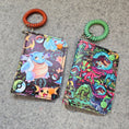 Load image into Gallery viewer, 2 vinyl bifold wallets with stretch bracelets laying on a dark gray surface.
