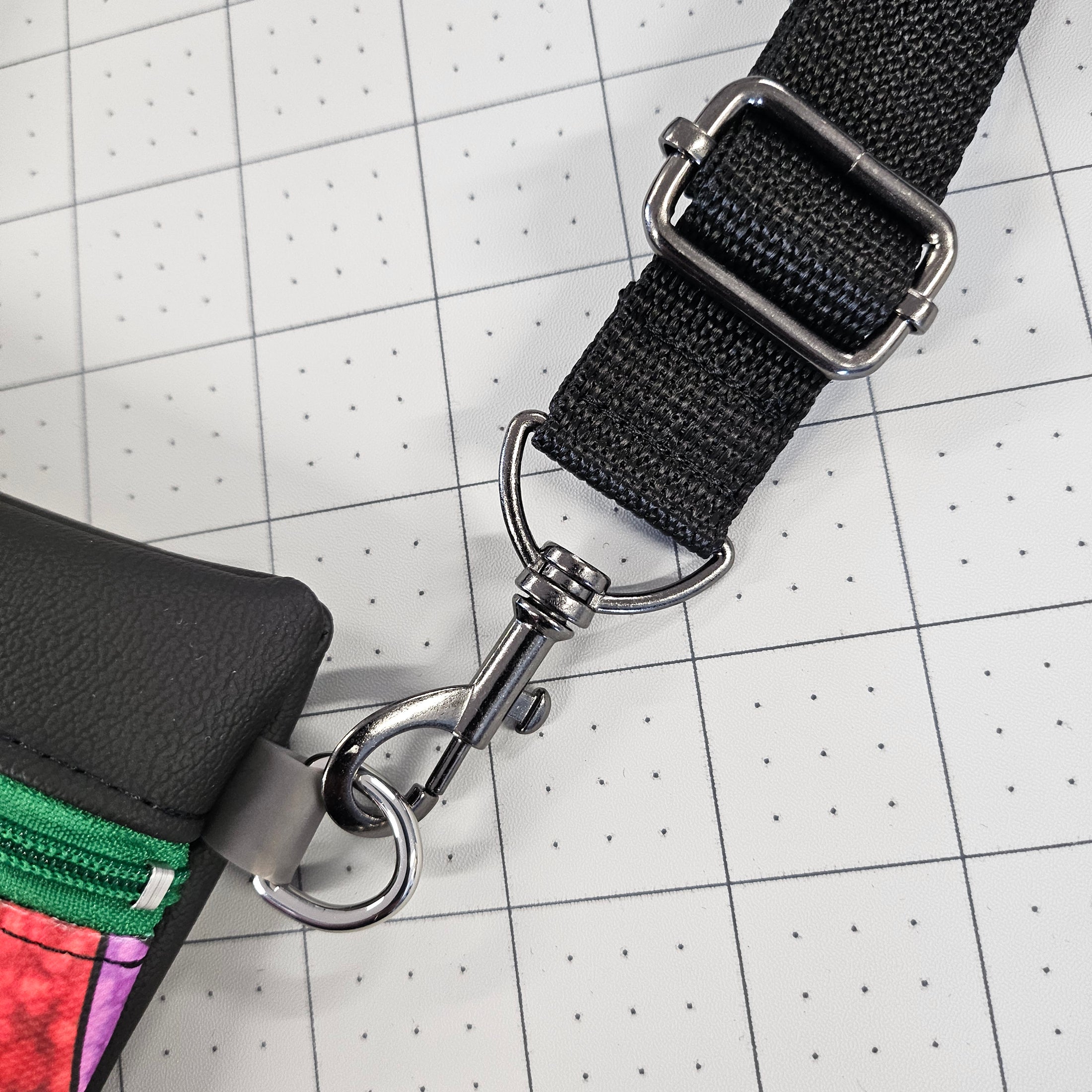 Black strap with metal buckle attached to a colorful pouch on a grid background
