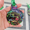 Load image into Gallery viewer, T Rex ornament with green ribbon held by a hand.
