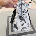 Load image into Gallery viewer, Hand holding a white clutch with black ink illustrations on a tiled floor background
