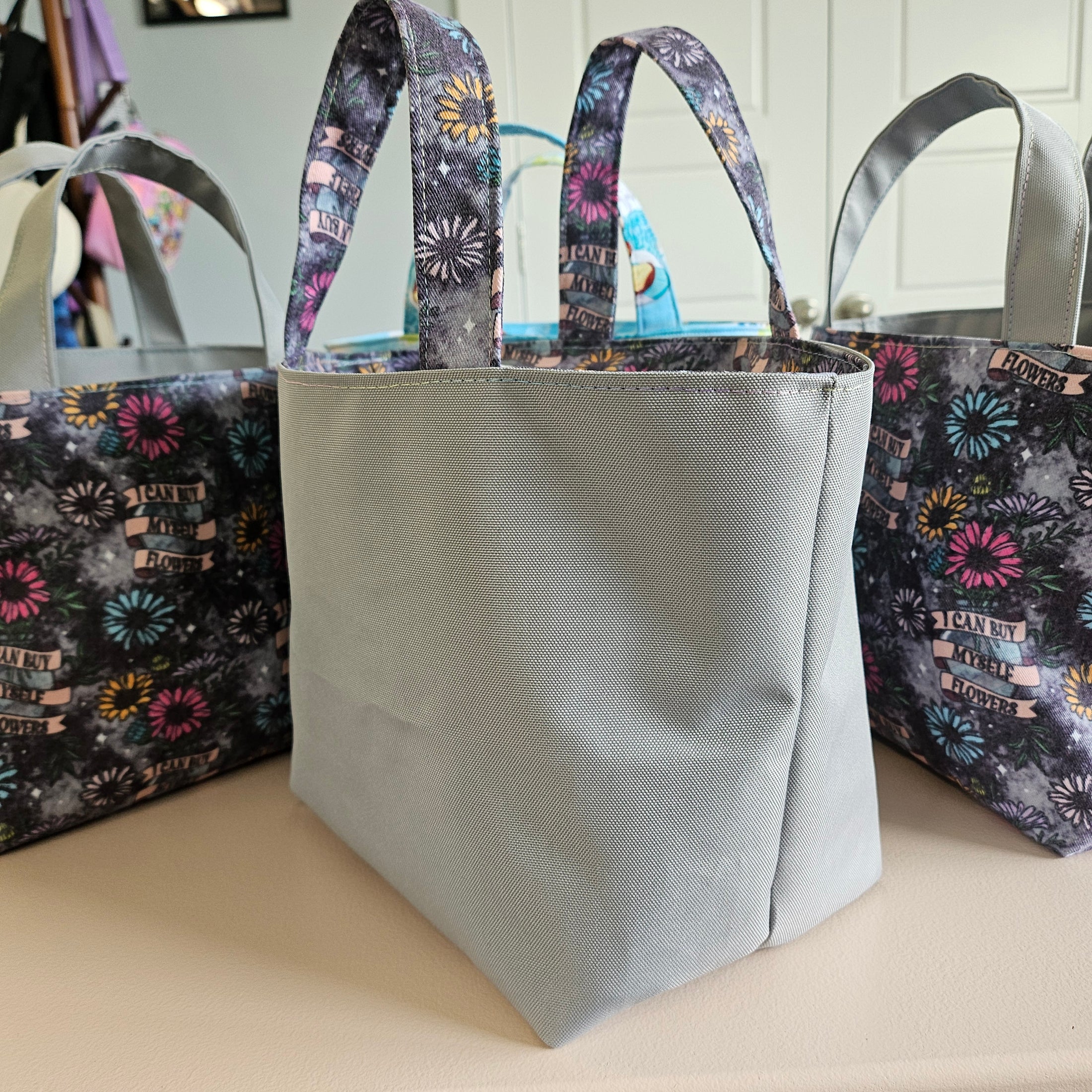 A light gray fabric tote bag with dark patterned handles and a floral print body, displayed on a table with two matching bags partially visible in the background.