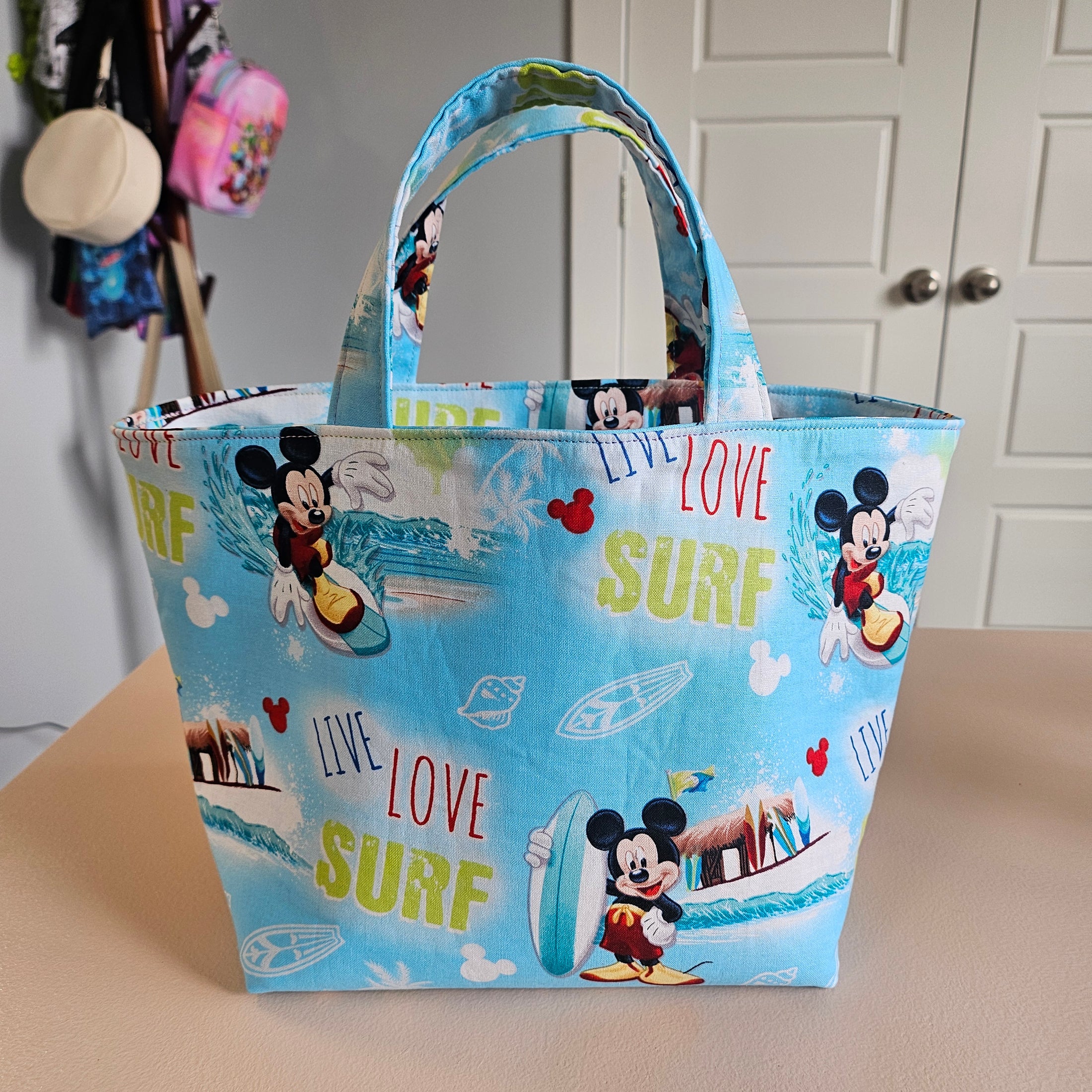 A light blue fabric tote bag with Mickey Mouse and Minnie Mouse prints, featuring 'LIFE LOVE SURF' text and surfboard motifs, shown hanging against a beige wall.